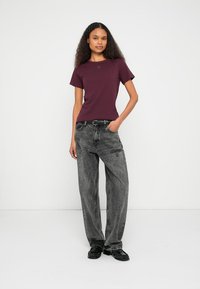 Vinrød T-skjorte med korte ermer og en liten logo, kombinert med baggy grå jeans. Sorte sko fullfører antrekket.