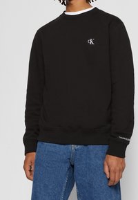 Felpa nera con scollatura a girocollo e polsini a coste. Presenta un piccolo logo CK bianco sul petto e il marchio Calvin Klein sulla manica.