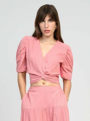 Femme portant un chemisier rose court à manches bouffantes et devant torsadé, porté avec une jupe taille haute assortie, se tenant debout les mains dans les poches.