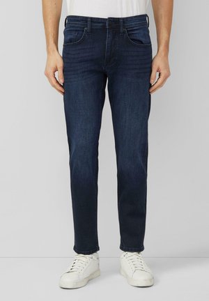 Mężczyzna w ciemnoniebieskich, dopasowanych jeansach i białych trampkach, stojący na jednolitym jasnym tle.