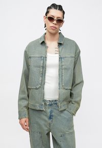 Τζιν μπουφάν με ξεβαμμένη μπλε απόχρωση, εμπρόσθιες τσέπες και γιακά. Φορεμένο πάνω από λευκή αμάνικη μπλούζα, συνδυασμένο με τζιν παντελόνια στο ίδιο χρώμα.