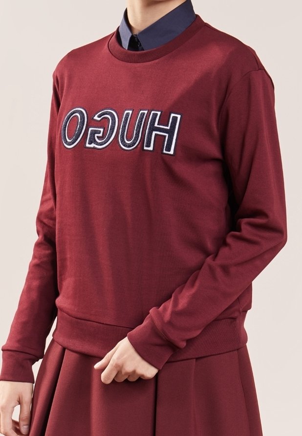 Personne portant un sweat-shirt rouge foncé avec « HUGO » écrit en lettres noires et blanches inversées, assorti à une jupe et une chemise bleu foncé à col.