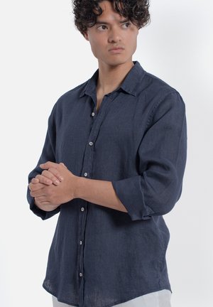 Giovane uomo con capelli ricci scuri che indossa una camicia blu navy abbottonata con le maniche arrotolate, con le mani giunte, che guarda di lato.