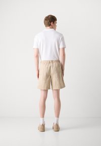 Beige bavlněné šortky s elastickým pasem a bočními kapsami, nošené s bílou košilí s krátkým rukávem a béžovými botami, zobrazené zezadu.