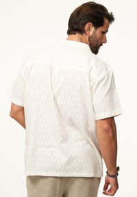 Kurzärmeliges weißes Hemd mit einem strukturierten geometrischen Muster, Kragen und geradem Saum. Der Stoff wirkt leicht und atmungsaktiv.