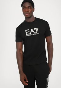 Sort bomulds T-shirt med en hvid "EA7 Emporio Armani" logo. Korte ærmer, klassisk crew neck design og en tætsiddende silhuet.