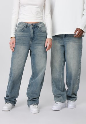 Δύο άτομα φορούν ανοιχτό μπλε, φαρδιά τζιν και λευκά αθλητικά παπούτσια, συνδυασμένα με λευκά μπλουζάκια μακρυμάνικα, στέκονται δίπλα δίπλα.