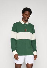 Zielona koszulka polo z długim rękawem, z kremowymi poziomymi paskami, wykonana z bawełny, z guzikowym kołnierzykiem i małym logo na klatce piersiowej.