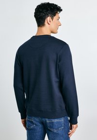 Man met kort krullend haar, gekleed in een effen navy sweatshirt en blauwe spijkerbroek, staand met zijn rug naar de witte achtergrond gekeerd.
