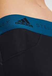 Czarne legginsy z szerokim morskim pasem w talii, na którym znajduje się czarne logo Adidas. Gładka tekstura i dopasowany krój z detalami w postaci szwów.