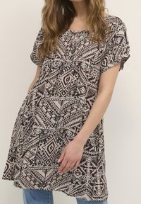 Kobieta nosząca luźną, krótką tunikę z geometrycznym czarnym i beżowym wzorem, założoną na niebieskie dżinsy.