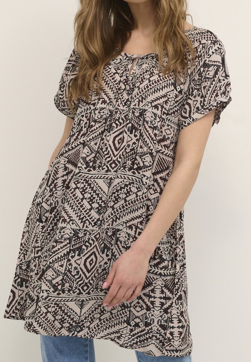 Kobieta nosząca luźną, krótką tunikę z geometrycznym czarnym i beżowym wzorem, założoną na niebieskie dżinsy.