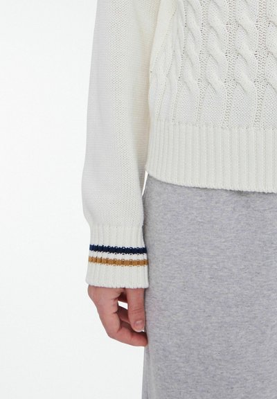 White knitted sweater with textured cable design, ribbed hem, and cuff featuring stripes in navy, gold, and white. Gray fabric beneath.