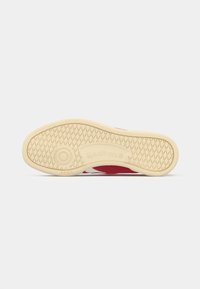 Suola di sneaker rossa con una superficie in gomma beige testurizzata. Presenta un logo circolare e scanalature per la trazione. Design minimale, adatto per un abbigliamento casual.