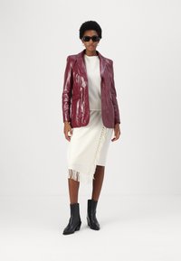 Blazer in pelle bordeaux, maglione crema e gonna bianca con frange. Stivaletti neri con tacco e occhiali da sole completano l'outfit. Texture liscia.