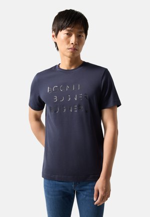 Mężczyzna w granatowej koszulce z krótkim rękawem z nadrukiem "BOGNER" trzy razy z przodu, w połączeniu z niebieskimi dżinsami, stojący na białym tle.