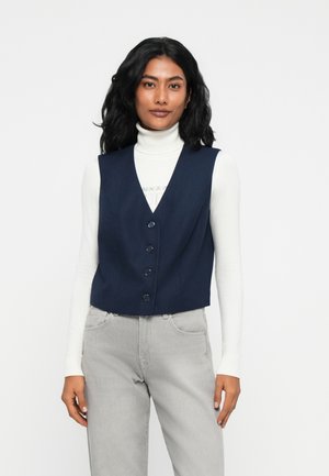 Kvinne med langt svart hår iført en hvit turtleneck, marineblå knappet vest, og lysegrå jeans, stående mot en hvit bakgrunn.