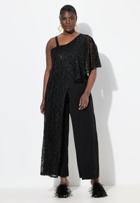 Top negro asimétrico con superposición de encaje, combinado con pantalones anchos negros. Presenta encaje texturizado, diseño de un solo hombro y tacones negros con plumas.