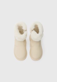 Bottines d’hiver beiges avec une épaisse doublure en fourrure blanche et des bouts arrondis, vues de dessus sur un fond uni.
