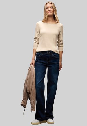 Beige Langarmshirt, dunkelblaue Jeans und eine hellbraune Wildlederjacke. Das Model trägt weiße Sneakers und steht vor einem grauen Hintergrund.
