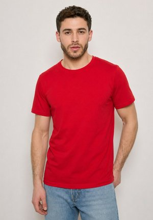 Jeune homme aux cheveux courts et bouclés portant un t-shirt rouge uni à manches courtes et un jean bleu clair, debout devant un fond neutre.