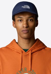 Navy baseball cap with embroidered white logo, worn with an orange hoodie featuring a circular graphic and drawstring detail.