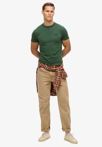 Groene t-shirt met korte mouwen, lichte stof, gecombineerd met tan broek en een rode geruite flanel over de taille geknoopt. Beige sneakers.