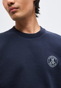 Νέος άνδρας που φοράει μπλε σκούφο φούτερ με λευκό κυκλικό λογότυπο που γράφει «FC MARC O'POLO STOCKHOLM 1967» στο στήθος.