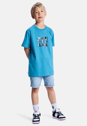 Jasnoniebieska bawełniana koszulka z kolorowym logo DC, zestawiona z jasnymi szortami dżinsowymi i czarnymi trampkami z czerwonymi akcentami.