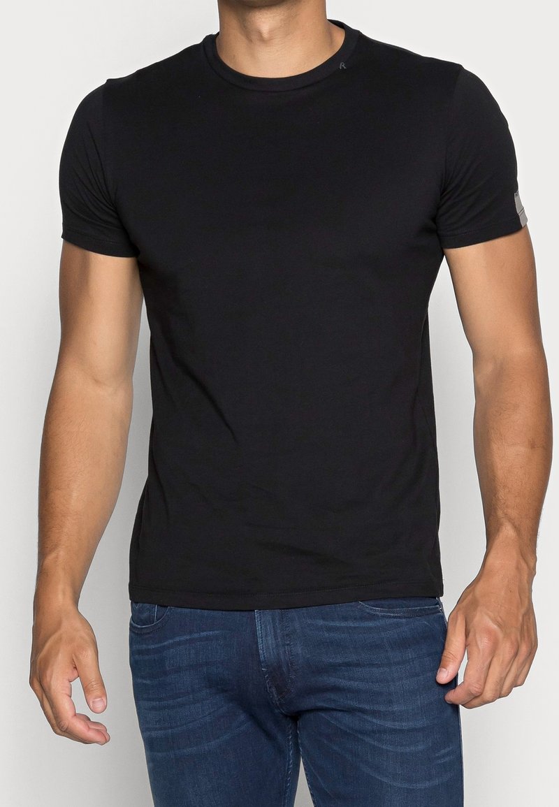 T-shirt noir à manches courtes en coton, présentant un col rond et une coupe décontractée, avec un discret logo sur la manche et une texture lisse.
