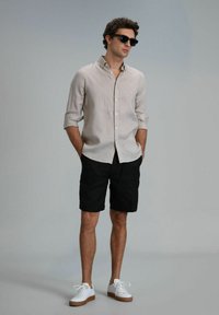 Lichtbeige linnen overhemd met opgerolde mouwen, gecombineerd met zwarte shorts en witte sneakers met bruine zolen. Zwarte zonnebril gedragen.