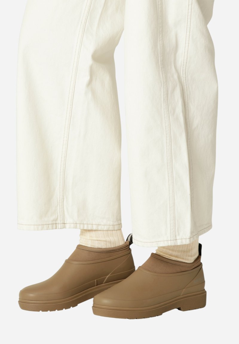 Brązowe gumowe botki za kostkę z teksturowaną podeszwą i brązowymi elastycznymi wstawkami, w połączeniu z kremowymi spodniami o szerokich nogawkach i jasnymi skarpetkami.