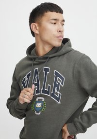 Oliven grøn hættetrøje med "YALE" trykt i fede hvide bogstaver. Har et rundt emblem med en bog og laurbærblade. Blød tekstur, hætte med snor.