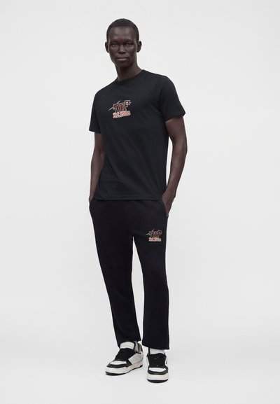 Homme portant un t-shirt noir et un pantalon avec un texte graphique rouge et blanc assorti, posant avec les mains dans les poches sur un fond uni.
