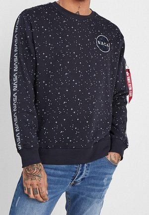 Czarna bluza z białym wzorem gwiazdek, logo NASA na piersi, napis NASA na rękawach, czerwona metka na lewej ręce, noszona z niebieskimi jeansami.