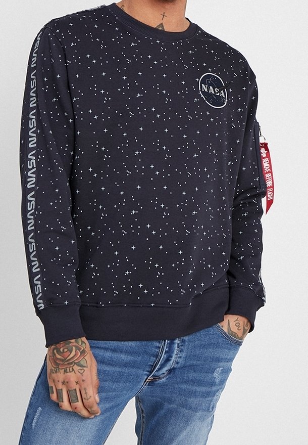 Sweat noir avec motif d'étoiles blanches, logo NASA sur la poitrine, texte NASA sur les manches, étiquette rouge sur le bras gauche, porté avec un jean bleu.