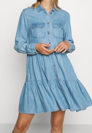 Woman wearing light blue denim button-up dress with long sleeves, two chest pockets, and a ruffled hem, hands adjusting front buttons.