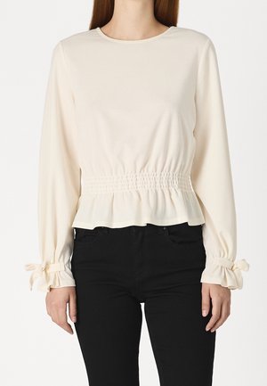 Frau trägt eine cremefarbene Langarmbluse mit geraffter Taille und gebundenen Manschetten, kombiniert mit schwarzen Hosen.