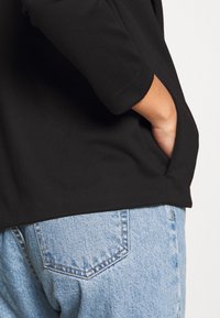 Svart tygtröja med trekvartsärmar, hand i fickan, kombinerad med ljusblå denimjeans som har en broderad bakficka.