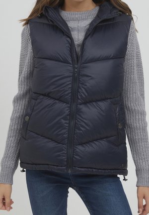 Kobieta w granatowej pikowanej kamizelce puchowej na jasnoszarym dzianinowym swetrze i niebieskich dżinsach, stojąca na jednolitym tle.