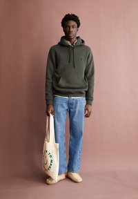 Olivgrüner Kapuzenpullover, hellbeiges Hemd darunter, blaue Jeans mit weitem Bein, cremefarbene Slipper und beige Tragetasche mit grünem Text.