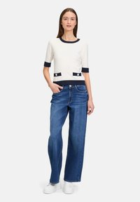 Gehaakte crèmekleurige top met marineblauwe rand, twee voorzakken met knopen. Loszittende lichtblauwe jeans, witte sneakers en een recht model.