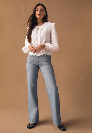 Kobieta stojąca na beżowym tle, ubrana w białą bluzkę z dużym kołnierzem, jasnoniebieskie dzwoniaste dżinsy oraz czarne baleriny z ćwiekami.