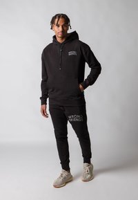 Set di felpa con cappuccio nera e pantaloni della tuta, con logo bianco "WRONG FRIENDS". Il materiale sembra morbido e strutturato. Sneakers chiare completano l'outfit.