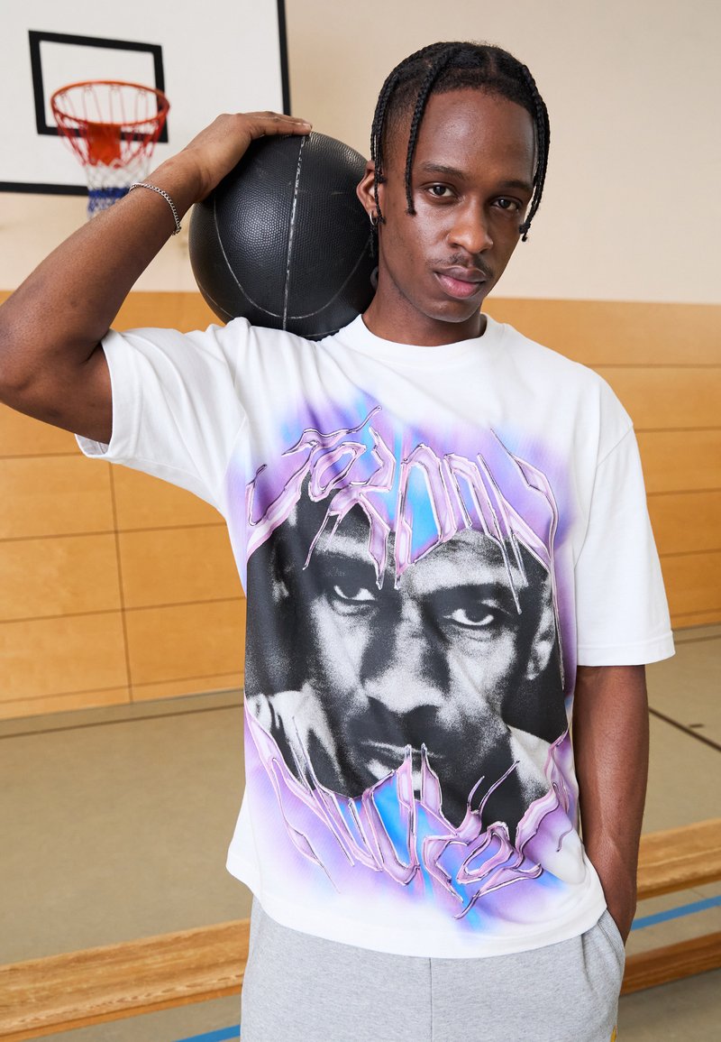 Hvit oversized T-skjorte med trykk av et ansikt i svart og lilla, med en teksturert basketball balansert på skulderen.