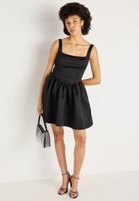 Melnas satīna kleitas ar kvadrātveida dekoltē, platām siksnām un izteiksmīgu svārku. Iezīmēta ar savelkamu jostu un gludu tekstūru. Melnas siksnainas kurpes un sudraba soma.