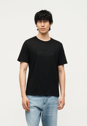 Młody mężczyzna z kręconymi czarnymi włosami, ubrany w czarny t-shirt "Originals" i jasnoniebieskie dżinsy, stojący na jednolitym białym tle.