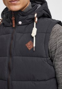 Chaleco negro acolchado con cuello alto y capucha, que presenta un parche de cuero con el logo, acentos de cuerda y cierre de cremallera.