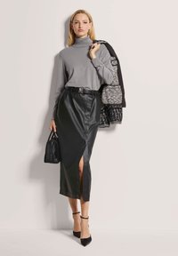 Grau Rollkragenpullover, schwarze Leder-Midirock mit seitlichem Schlitz, schwarzer Gürtel und schwarze Handtasche; Modell trägt schwarze High Heels und hält eine Jacke.