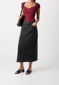 Femme portant un haut côtelé bordeaux à manches courtes, une longue jupe en jean noire taille haute avec un ourlet brut, des sandales à talons noires, et portant un sac à bandoulière noir.